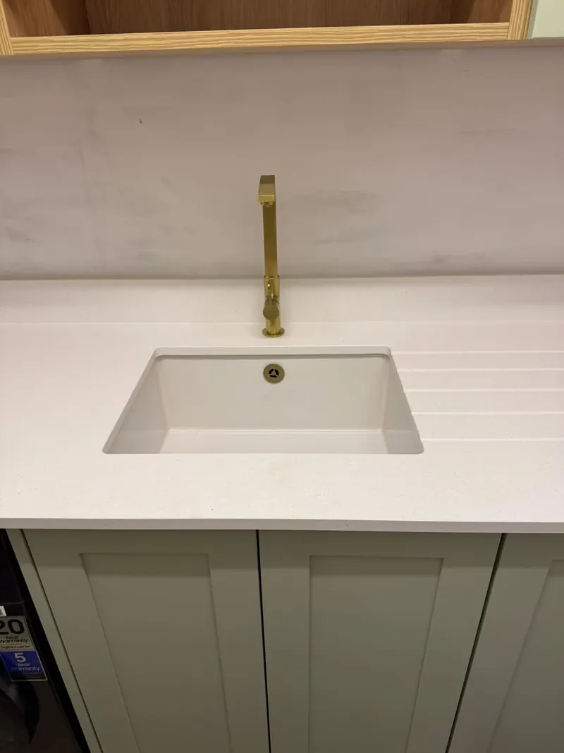 Undermount kitchen sink with a brushed brass tap in a white worktop with drainer grooves, above sage green cupboards